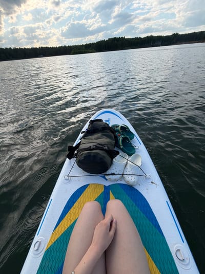 The image depicts a person sitting on a paddleboard in a body of water. The board is white with blue, green, and yellow accents, and has a dark bag, a pair of green Crocs, and a pair of white Crocs secured to the front. The person has their legs bent and their hands resting on their legs. The water is dark and rippled, and the shoreline with trees is visible in the distance. The sky is partially cloudy. It's impossible to verify the location as Selychka, Russia from the image.