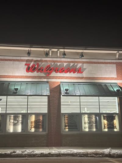 The image displays the exterior of a Walgreens store building at night, seen from a slightly elevated perspective. A prominent red "Walgreens" sign, oriented vertically on the building's facade, glows brightly, casting a soft shadow on the lighter brick and paneling behind it. Below the sign, a series of five wall-mounted spotlights illuminate the wall.
To the left of the building is a dark, undefined area, signifying the nighttime setting. To the right of the vertical sign are large windows, reflecting vertical light fixtures from the interior and displaying framed historical-looking images or advertisements within the store. Above and below these main window sections, there are dark, possibly metallic, panels, one with a small light fixture. The building's architecture features a combination of warm-toned brickwork and lighter, possibly stone or concrete, panels. On the far right edge of the image, a vertical section of a building or column appears textured and covered in a white, granular substance, strongly suggesting the presence of snow or ice.
The scene depicts a commercial establishment, specifically a Walgreens in Leominster, United States, well-lit against the dark night, possibly during winter due to the icy appearance on the right. No people or specific activities are visible, focusing instead on the illuminated building and its features.