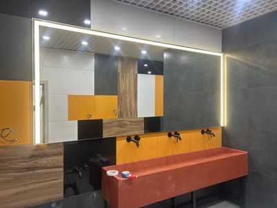 The image shows a modern bathroom with a long, orange-red concrete sink. Two black faucets are mounted on the wall above the sink. The wall behind the sink is a mix of white, orange, and wood-patterned tiles. A large, illuminated mirror stretches across the top of the sink area, reflecting the tiled wall and the ceiling, which has a grid pattern. To the right of the sink, the wall is covered in dark grey tiles. On the sink, there is a roll of white tape and a small bottle of red liquid. The scene appears to be located in the city of Druzhny, Russia.