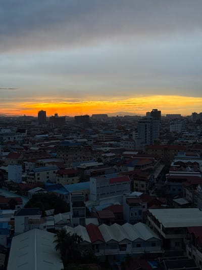 This is an aerial photograph of a city, likely Khan Mean Chey, Cambodia, during sunset. The image is oriented vertically, with the sky occupying the left half and the cityscape on the right.

The sky is filled with dramatic sunset colors, transitioning from a pale gray at the top left to vibrant hues of orange and yellow near the horizon. Wispy clouds are visible, catching the warm light of the setting sun.

The right half of the image displays a dense urban landscape composed of numerous buildings with varying architectural styles and roof colors, including red, blue, and white. Many of the buildings appear to be residential or mixed-use, with some taller structures interspersed among smaller ones. The overall impression is of a bustling city viewed from above.

The time of day is clearly sunset, indicated by the intense coloration of the sky and the shadows cast across the buildings. The weather appears clear, allowing for an unobstructed view of the sunset. There are no people visible, and the primary subject is the urban scene illuminated by the sunset. No visible text is present in the image.