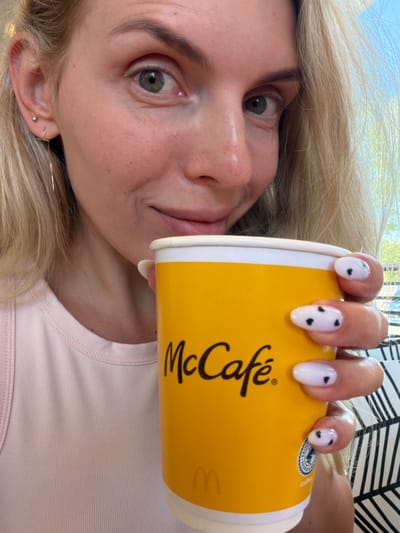 A close-up, selfie-style shot shows a fair-skinned woman with blonde hair and green eyes holding a yellow McCafé cup. She is wearing a light pink tank top. Her nails are painted white with black polka dots. The background is blurred, but appears to be an outdoor setting with sunlight filtering through trees and a portion of a black and white striped pattern. The image was taken in the city of Húmera-somosaguas-prado Del Rey, Spain.