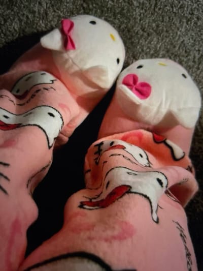 A high-angle, close-up photograph shows two feet, encased in plush pink footed pajamas or slippers, resting on a textured grey carpet. The left foot's covering features a white Hello Kitty head with black dot eyes, a yellow oval nose, and a small pink bow on its left ear. The right foot's covering displays a similar white Hello Kitty head with black dot eyes and a yellow oval nose, but with a larger, prominent pink bow positioned over its mouth area. The pink fabric extending from the foot coverings is patterned with multiple cartoon figures resembling white ducks or birds, outlined in black, with some featuring red accents. A dark shadowed area is visible between and beneath the feet. The scene is located in Arthès, France.