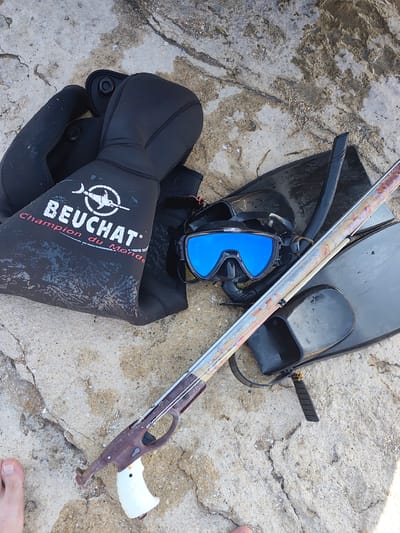 A spearfishing equipment set lies on a textured, rocky surface with some moisture present. The gear includes a black wetsuit with the brand "BEUCHAT" visible, a pair of black fins, a snorkel, a mask with bright blue lenses, and a speargun. The speargun is metallic with a brown handle and a white grip. A human foot, partially visible on the bottom left, suggests a person is nearby. The environment appears to be outdoors, possibly near a body of water, given the nature of the equipment. No specific location in Saiyn Şapağatov, Kazakhstan is indicated by the visual evidence.