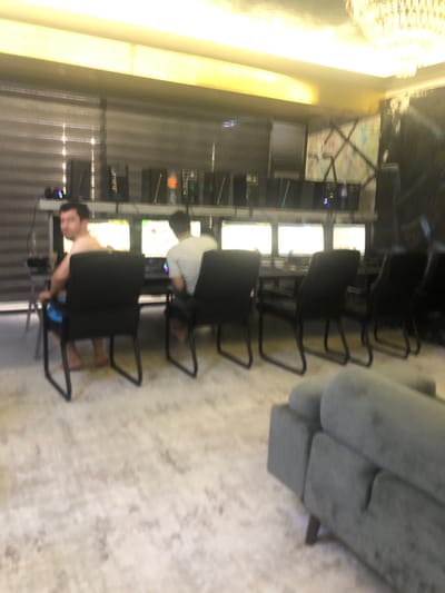 In the city of Khujakent, Uzbekistan, a dimly lit room features a long table with multiple computer monitors displaying bright screens. Two men are seated at the table, facing the monitors. One man, shirtless and wearing blue shorts, is seated in a black chair on the left, looking back over his shoulder. To his right, another man in a striped t-shirt and shorts is seated, facing away from the camera. Several black computer chairs are positioned around the table. The room is furnished with a large, gray sectional sofa in the foreground. The walls are decorated with dark paneling and a framed artwork. A large, ornate chandelier hangs from the ceiling. The lighting is predominantly from the computer screens and overhead fixtures, creating a yellow glow.