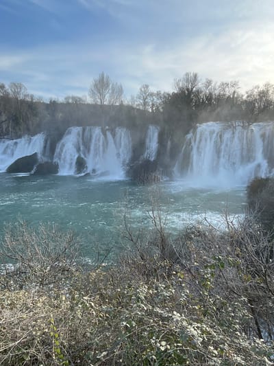 The image displays the Kravica Waterfalls located in Zvirići, Bosnia and Herzegovina. The scene is dominated by multiple wide, natural cascades of white water plunging into a broad, turbulent body of turquoise-colored water below. Significant mist rises from the impact zones of the falling water. The midground and background feature a dense line of mostly bare deciduous trees and brush, suggesting a cool season. The foreground contains various leafless branches and sparse green foliage belonging to low-lying bushes and shrubs. The sky above is light blue with scattered wispy white clouds. No human subjects, animals, or artificial structures are visible. The primary action is the continuous flow and fall of water over rock formations.