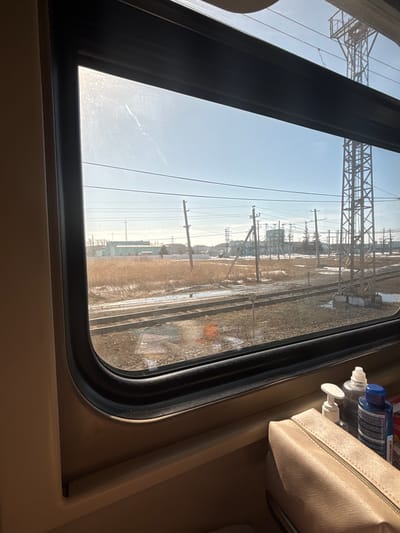 The image provides a view from inside a train, looking out a window onto a railway environment in the city of Kalachinsk, Russia. The scene is illuminated by bright daylight under a clear, light blue sky.
Inside the train, a section of a beige interior wall and the dark-framed window are visible. On the window's lower ledge, a light beige/peach-colored zippered pouch is placed, behind which stand three bottles: a white pump-dispenser bottle, a clear plastic bottle with a silver cap, and a blue plastic bottle with a blue cap. No individuals are present in the frame.
Outside the train, multiple parallel railway tracks extend across the mid-ground. The ground surrounding the tracks is covered with dormant, light brown vegetation and scattered patches of snow or ice, indicating a late winter or early spring setting. Numerous utility poles, some wooden and others metal, support a network of overhead wires that crisscross the sky. A tall, dark metal lattice tower, likely a transmission or communication structure, is prominent on the right side of the view. In the background, several industrial or commercial buildings with flat roofs are discernible, one of which features a distinct turquoise or light green facade. Further utility infrastructure and sparse, bare trees are also present in the distance.