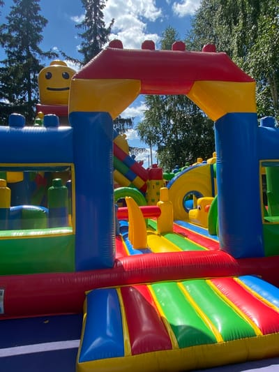 An inflatable playground shaped like a Lego castle sits outdoors on a sunny day. The structure is primarily blue, red, and yellow, with large Lego-like blocks and a smiling Lego head at the top. It features slides, tunnels, and various play areas. The playground is situated on a dark blue surface, possibly a mat. In the background, tall green trees and a bright blue sky with scattered white clouds are visible. The scene appears to be in a park or outdoor recreational area.