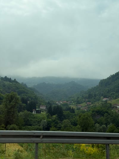 The image depicts a hazy, mountainous landscape in Lašva, Bosnia and Herzegovina. The scene is captured from a roadside vantage point, indicated by a metal guardrail in the foreground on the right side of the frame.

The left side of the image is dominated by a soft, diffused sky, suggesting overcast or foggy weather. As the view moves to the right, a dense, green forest blankets the rolling hills. The trees are lush and full, indicating a temperate climate, likely during spring or summer. Scattered within the treeline and on cleared patches of the hillsides are small houses with red-tiled roofs, hinting at a rural or semi-rural settlement. The perspective suggests that these houses are built on slopes, following the contours of the terrain.

Further to the right, beyond the forested slopes, a grassy embankment can be seen, with hints of yellow wildflowers. The overall atmosphere is serene and natural, with a sense of peacefulness. The haziness in the distance creates a layered effect, adding depth to the landscape. There is no visible text in the image. The time of day appears to be daytime, though the overcast conditions make it difficult to determine the exact hour. The weather seems to be damp or misty, contributing to the soft focus and diffused light.