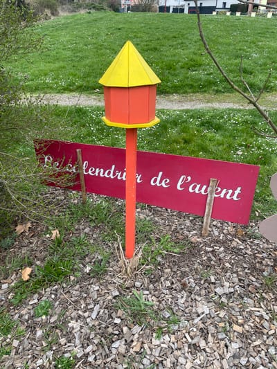 The image depicts an outdoor environment in Holtzheim, France. Central to the foreground is an erected hexagonal structure, approximately one meter in height, featuring an orange body, a yellow conical roof, and a yellow circular base. This structure is mounted on a cylindrical orange wooden pole, secured in a ground area covered with light-colored wood chips, green grass, and dried vegetative matter. Immediately behind and partially to the left of this pole, a horizontal rectangular sign, painted red with white serif lettering, displays the text "Le calendrier de l'avent." The sign is supported by two unpainted wooden stakes embedded in the wood-chipped and grassy ground. To the left of the sign, a green deciduous bush is present. On the right, bare tree branches extend into the frame. The midground features an expansive green lawn containing numerous small white flowers. A narrow, light-colored dirt path traverses this lawn. In the distant background, residential buildings with dark roofs and light walls are visible among sparse trees and foliage under an indistinct sky. No human figures or active subjects are depicted within the scene.