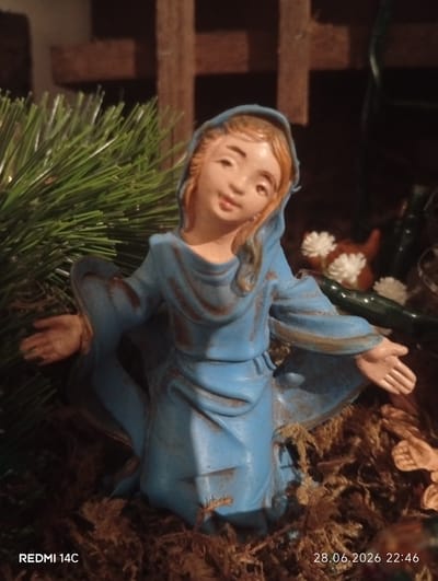 A close-up, low-angle shot captures a small, ceramic nativity figurine of a woman in a blue robe. The figurine is positioned in the center of the frame, with its arms outstretched and palms facing upward. The woman has light brown hair and fair skin, with a gentle expression on her face. She is kneeling on a bed of dried moss, which surrounds her and fills the foreground. Behind the figurine, a blurred background features a wooden cross structure and some greenery, resembling a Christmas nativity scene. The lighting is dim, casting shadows and highlighting the texture of the moss and the figurine's robe. The bottom left corner of the image displays the text "REDMI 14C," and the bottom right corner shows "28.06.2026 22:46". The scene does not provide any specific information about the city of Civitanova Marche, Italy.