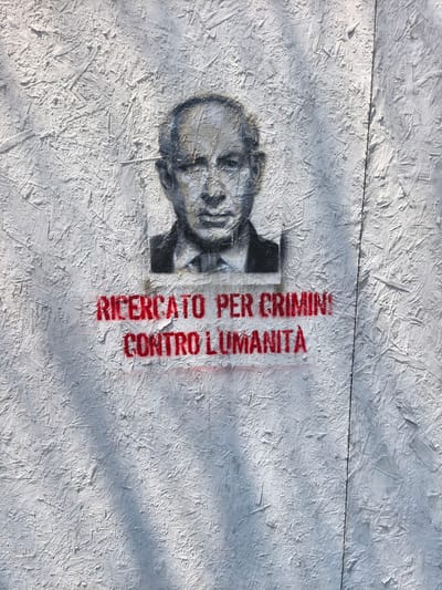 This is a photograph of a stencil graffiti art piece on a wall made of OSB (Oriented Strand Board). The graffiti depicts a portrait of a man in black and white, rendered in a somewhat rough style with visible spray paint texture. The man is wearing a suit and tie and appears to be bald with glasses. Below the portrait, in bright red stencil lettering, are the words "RICERCATO PER CRIMINI CONTRO L'UMANITÀ", which translates from Italian to "WANTED FOR CRIMES AGAINST HUMANITY".

The setting appears to be outdoors, as evidenced by the dappled sunlight casting shadows across the OSB surface and the graffiti. The OSB material itself, with its visible wood fibers and textured surface, suggests an unfinished or temporary structure, possibly a construction site hoarding or a temporary barrier. The background is largely out of focus, emphasizing the graffiti. The overall mood is serious and accusatory, conveyed by the "wanted" poster style and the grave accusation of "crimes against humanity." The lighting suggests it is daytime, likely with clear skies given the sharp shadows. The location context provided is Padua, Italy, which implies the graffiti carries a message relevant to that area or a broader political commentary. The red paint of the lettering appears to have bled slightly in places, adding to the raw aesthetic of the stencil art.