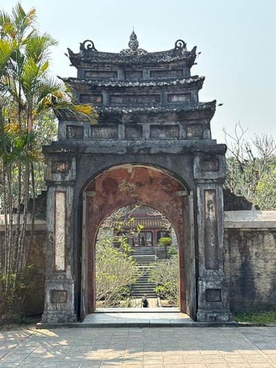 The image depicts an ornate stone archway, with birds perched atop its roof, in Kim Long Ward, Vietnam. The archway leads to a courtyard with a building and stairs. The archway's structure is aged and textured with decay. Palm trees and other greenery are visible near the archway. A paved walkway is on the right of the frame.