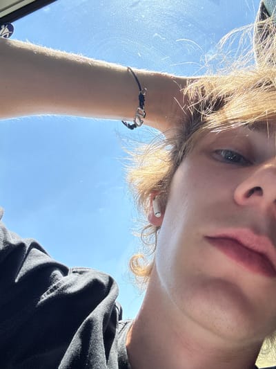 The image is a close-up, low-angle selfie taken outdoors, with a bright blue sky dominating the background. The subject, a young person with blonde hair, is looking directly into the camera. They are wearing a black t-shirt and have a white wireless earbud in their right ear. A bracelet with a silver charm is visible on their left forearm. Sunlight is strongly illuminating the scene, creating highlights on the subject's hair and skin. The image is framed by what appears to be the edge of a window or windshield, with some smudges and reflections visible on its surface. A small circular blue and white decal is affixed to the upper left corner of this surface. Given the context, the selfie was likely taken in the vicinity of Cadeo, Italy, potentially from within a vehicle given the window-like framing.