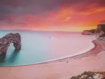 The image depicts a picturesque coastal landscape at sunset. A prominent rock arch formation, likely a sea stack, stands in the turquoise water on the left side of the frame. A sandy beach curves along the shoreline, leading to gentle waves lapping the shore. In the distance, rolling hills and cliffs form the coastline. The sky is ablaze with warm hues of orange, pink, and purple, indicative of a dramatic sunset. There are no discernable people or specific landmarks that would associate this scene with Moelv, Norway.
