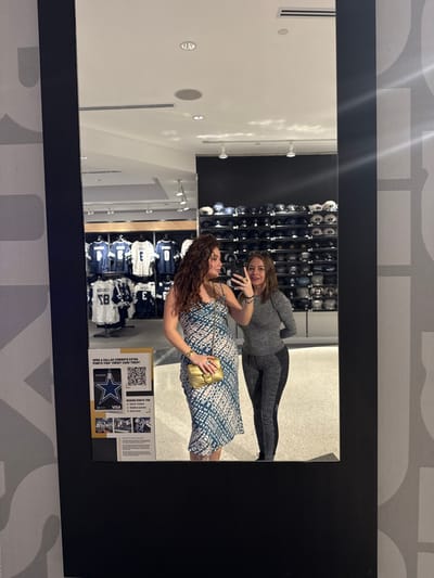 Two women are posing for a picture in a mirror inside a store. One woman has curly brown hair and is wearing a blue and white dress with a gold purse. The other woman has short brown hair and is wearing a gray top and black leggings. The mirror reflects a wall of Dallas Cowboys apparel, including hats and jerseys. A promotional sign with a QR code and the Dallas Cowboys logo is visible on the right side of the mirror.