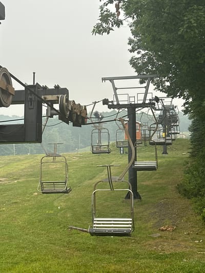 The image depicts a ski lift on a grassy slope under an overcast sky. The ski lift structure is a metal pole supporting a cable with several empty chairs attached. The chairs are made of metal with wooden slats. In the background, trees and hills are visible, suggesting a rural or mountainous setting. There is a small red building with windows to the left of the ski lift. The ground is covered in green grass with scattered small white flowers. The overall scene suggests a quiet, possibly off-season moment for the ski resort.