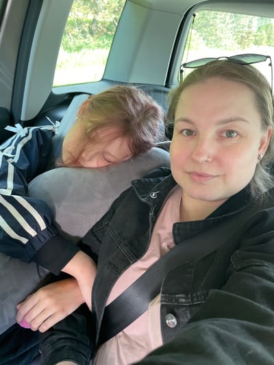 The image shows a selfie taken inside a vehicle, likely a car. A woman with fair skin and light brown hair, wearing a black jacket over a pink shirt and a seatbelt, is visible in the upper part of the frame. Sunglasses are placed on top of her head. A young girl with light brown hair is resting on a grey pillow next to her. The girl is wearing a navy blue jacket with white stripes and white bows. Through the car window, green trees are visible. The location is stated as Средний Постол, Russia, however, no elements in the picture can confirm this.