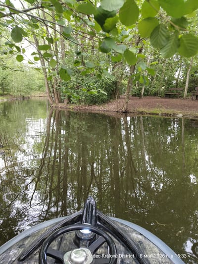 A photograph taken from the vantage point of a boat traveling on a body of water, potentially a river or canal, near Chlumec nad Cidlinou, Czechia. The boat's steering wheel and front deck are visible in the foreground. The water reflects the surrounding trees, mostly deciduous, with green foliage. The water is a murky green-brown color. The banks of the waterway are lined with trees and vegetation, with a bench visible on the right bank. The date and location are stamped on the bottom of the image: "Hradec Králové District | 8 май 2026 г. в 15:33 ч."