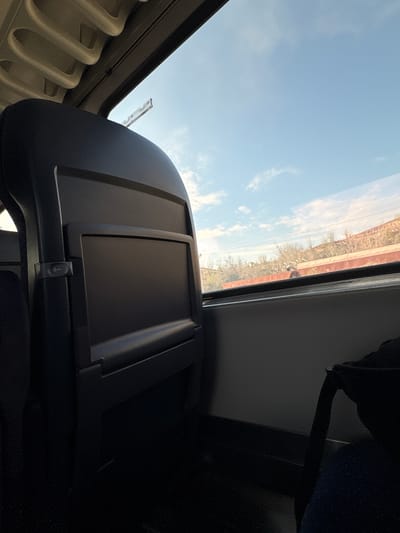 The image is taken from inside a vehicle, likely a train or bus, looking up at the sky and the passing landscape. The interior elements, such as a seatback tray, are in the foreground, while the sky with clouds and the blurred landscape are visible through the window. The location is not determinable from the image content.