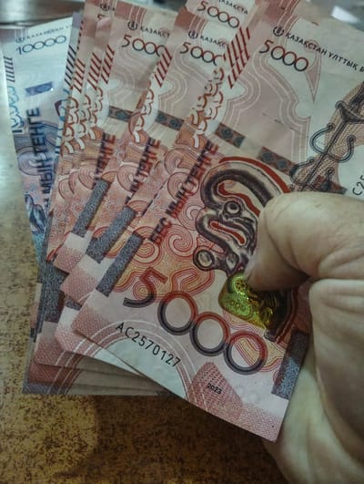 This media file displays a close-up, top-down view of a hand holding a fanned stack of Kazakhstani Tenge banknotes. The banknotes are predominantly pink and white with intricate patterns. Several denominations are visible, including 10,000 Tenge and multiple 5,000 Tenge notes.
The hand, partially obscuring some of the bills, is gently pressing on a holographic security feature on one of the 5,000 Tenge notes. Visible text on the banknotes includes "ҚАЗАҚСТАН" (Kazakhstan), currency denominations like "10000" and "5000", and serial numbers such as "AC2570127". The year "2023" is also visible on one of the notes.
The background consists of a textured surface, likely a table, with a mottled brown and yellow pattern. The lighting suggests an indoor setting, with no specific time of day or weather conditions indicated. The scene is focused on the display of money, possibly signifying a transaction, wealth, or the economy of Kazakhstan. The overall tone is neutral, focusing on the objects and their details. The location context provided suggests the images are related to Besagash, Kazakhstan, which aligns with the currency shown.