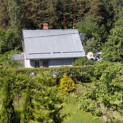 This is an aerial, top-down photograph of a residential property surrounded by dense forest and a well-maintained lawn. The house is a single-story structure with a grey, corrugated metal roof. A brick chimney is visible on the left side of the roof. Two satellite dishes are mounted on the roof; one is white with a rusty dish, and the other is grey and appears newer. There are visible power lines running across the frame.
The surrounding vegetation is lush and varied, with tall pine trees on the left and a mix of deciduous trees and evergreen shrubs on the right. A manicured lawn occupies the upper right portion of the image. The scene suggests a rural or semi-rural setting, consistent with the provided location context of Skuķīši, Latvia.
The time of day appears to be daytime, with strong sunlight casting distinct shadows, particularly on the lawn. The weather seems to be clear and sunny, conducive to outdoor activities or simply enjoying the natural surroundings. There is no activity or event clearly depicted; the image appears to be a static scene of the property.
Visible text on the grey satellite dish reads "UNISAT". No other text is discernible. The overall impression is one of a peaceful, secluded home nestled within a natural environment.