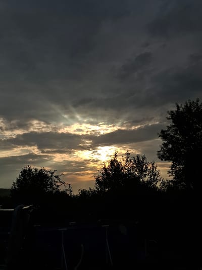 The image shows a dramatic sky with dark, heavy clouds, interspersed with rays of sunlight breaking through. The sun appears to be setting or has recently set, casting a warm, golden glow on the clouds. Silhouetted against the sky are the dark shapes of trees, including a large tree on the right and a cluster of trees in the middle ground. In the foreground, a dark, indistinct object, possibly an above-ground swimming pool, is visible with a blanket draped over its side. The scene suggests an outdoor setting at dusk. The location is identified as Sosnovka, Russia.