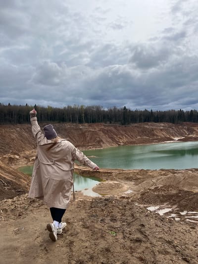 The image depicts a person standing on a dirt surface at the edge of a quarry filled with greenish water. The person is wearing a long beige trench coat, black pants, white socks, and white sneakers, and has a gray beanie on their head. Their arms are outstretched. The environment includes exposed dirt cliffs around the quarry, a treeline in the background, and a cloudy sky. The image has been rotated. The location is Ivlevo, Russia.