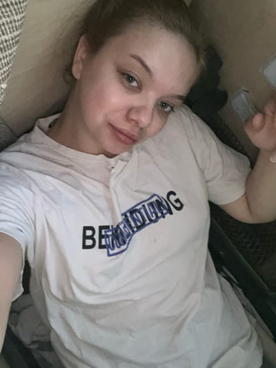 The image is a close-up, overhead shot of a young woman lying down, seemingly on a bed or sofa, looking towards the camera. She has light skin, light-colored eyes, and her hair is pulled back. She is wearing a white t-shirt with blue and black text on it. The text appears to read "BEAUTIFULLY". The background is dimly lit and shows what appears to be furniture and possibly bedding. The woman's expression is relaxed, and she has a slight smile. The lighting suggests it could be daytime, possibly indoors, given the lack of natural light cues. There are no clear indicators of the weather or specific location beyond the provided context of Gorodets, Russia.