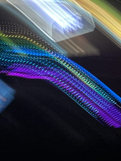 The image is an abstract close-up of a moving object, likely a light installation or a structure with lights, creating streaks of color and blurred patterns against a dark background. The colors are predominantly purple, blue, green, yellow, and white, appearing in diagonal lines and fragmented shapes due to motion blur. The texture of the background suggests a dark, possibly outdoor environment. There are no discernible people or specific landmarks to identify a precise location within Stallings, United States of America. The image captures a dynamic, light-based phenomenon rather than a static scene.