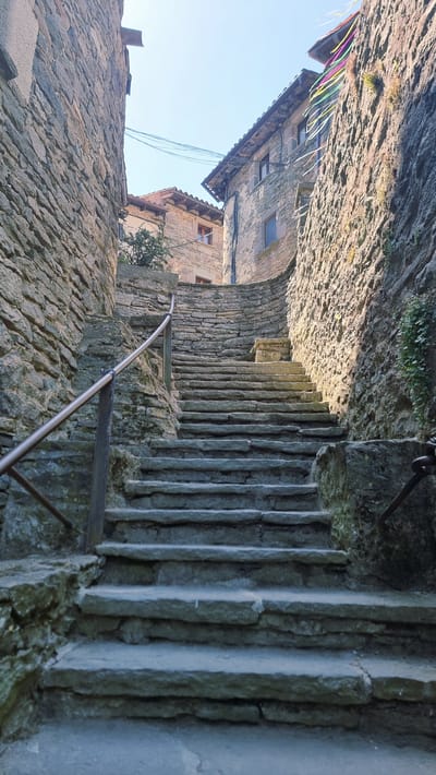 This is a photograph of an old stone staircase in the medieval village of Rupit i Pruit, Catalonia, Spain. The staircase is made of uneven, weathered stone steps and is flanked by rustic stone buildings. A metal handrail runs along the left side of the stairs. The buildings have stone walls and tile roofs, and the overall impression is one of antiquity and a historic European setting. The sky is visible at the top of the image, and there are some overhead wires and colorful ribbons strung between buildings. The lighting suggests it is daytime, with sunlight casting shadows on the scene.