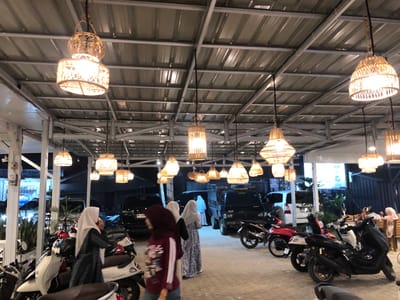This media file captures an outdoor seating area at night, likely a restaurant or cafe, in Blangbitra, Indonesia. The scene is illuminated by numerous woven pendant lights hanging from a corrugated metal roof supported by a metal frame. The lights create a warm, inviting ambiance.
Several people are present, mostly women wearing hijabs and modest clothing. In the foreground, a woman in a dark patterned dress and hijab is the most prominent subject. To her left, a woman in a lighter patterned dress and hijab stands near a white scooter. Further into the background, other individuals are visible, some seated at tables, while others are standing or walking.
A variety of motorcycles and scooters are parked in the area, suggesting it might be a popular spot for commuters or a place where people arrive on two-wheeled vehicles. The vehicles are of different colors and models, including a white "Scoopy" model. Black cars and a small truck are also visible in the background, parked beyond the seating area.
The overall setting appears to be an open-air establishment, with a concrete floor and partially visible walls or structures. The time of day is clearly night, with artificial lighting being the primary source of illumination. There are no immediate cues about the weather, but the open setting suggests it is likely pleasant.
No legible text is visible in the image. The mood seems relaxed and social, characteristic of an evening gathering at a local eatery.