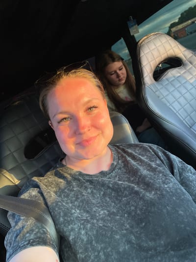 Two women are sitting in a car. The woman in the foreground is looking at the camera, smiling, with sunglasses on her head. The woman in the background is looking down and to her left. The car appears to be parked in an outdoor lot, possibly a rest stop or parking area. The time of day appears to be late afternoon or early evening, with the sun casting long shadows and a warm light. The location is not discernible within the city of Sovkhozny, Russia.