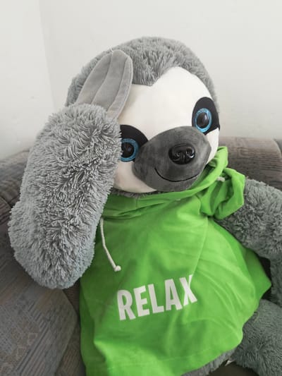 A plush toy sloth is positioned on its side, with its head towards the lower left of the frame. The sloth is predominantly gray with fluffy fur, white facial markings, and large, round blue eyes with black pupils. It has a dark gray snout with a black button nose and a faint smile line. The sloth is wearing a bright green hooded garment that drapes over its body and extends upwards and to the right. White text, "RELAX," is prominently displayed on the green fabric in a bold, sans-serif font. The sloth's paws are visible, with one appearing to be resting near its head.
The background consists of a textured surface, possibly a sofa or chair, with a striped pattern in shades of gray and beige. To the left and behind the sloth, a plain white wall is visible. The lighting appears to be natural, suggesting daytime, and there are no indicators of weather conditions. The overall mood evoked by the plush toy and the text on its garment is one of comfort and relaxation. The scene is likely indoors, given the presence of furniture and a wall. The location context provided is Oberviechtach, Germany.