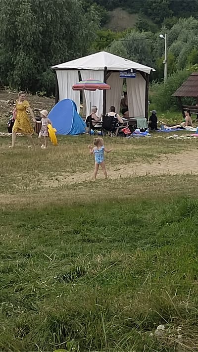 This is an outdoor scene, likely a beach or park, in Ramon, Russia. In the foreground, tall green grass dominates the lower half of the image. In the mid-ground, a young girl in a blue swimsuit is standing and appears to be dancing or playing. To the left, a woman in a yellow dress is holding the hand of a smaller child in a floral dress who is carrying a yellow ring. Behind them, a blue tent is set up. Further back, a white gazebo with draped curtains provides shade for several people, some of whom are seated. One person is in a wheelchair. To the right of the gazebo, a smaller wooden structure with a brown roof is visible. Several other individuals are seen lounging on the ground in the background. The environment appears to be a sunny day with trees and a hill in the distance.
