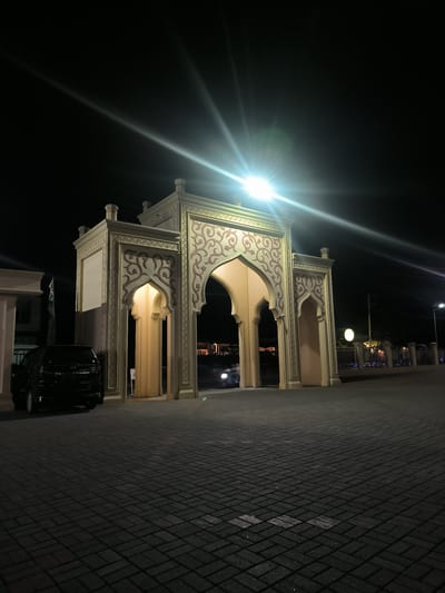 The image captures a grand, ornate gateway structure at night, illuminated by bright artificial lights that create prominent lens flares. The architecture of the gateway features multiple arches, pillars, and intricate swirling patterns in a reddish-brown hue against a cream or beige background, characteristic of Islamic design. The main gate appears to have a large central arch flanked by smaller arched entrances.
To the left of the gateway, a black SUV is parked, with its rear visible. A license plate, "DL 1704 JK," is clearly legible on the vehicle. Through the archways, the headlights of other vehicles are visible, suggesting activity and passage through the gate. The ground in the foreground is paved with dark, rectangular blocks, showing slight reflections from the surrounding lights.
In the background, beyond the gateway, dimly lit structures and a fence with decorative blue lights can be seen. The overall scene is set at night, with the sky appearing completely dark. There is no visible activity involving people, but the presence of vehicles indicates movement and functionality of the gateway, likely serving as an entrance or thoroughfare. The architectural style points to a cultural context common in regions like Indonesia, consistent with the provided location context of Mee Tanjong Usi.