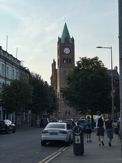The image shows a street scene in the city of Derry/Londonderry, United Kingdom, with a prominent clock tower, likely St. Columb's Cathedral, dominating the background. The sky is partly cloudy and appears to be late afternoon or early evening.

In the foreground, a paved street runs along the right side of the image, with double yellow lines marking the edge of the carriageway. Several cars are parked on the street, including a white Audi with the license plate GU62 XOW. A pedestrian sidewalk is to the right of the street, with three people walking away from the camera. The person in the middle is wearing a white t-shirt and a backpack, while the person on the right has pink hair and is wearing a dark top and shorts. A young person wearing a blue t-shirt and shorts stands near a trash can.

On the left side of the street, there are buildings with multiple windows and an architectural style that suggests they are older commercial or residential properties. Trees with dense foliage are planted along the sidewalk in front of these buildings. The clock tower, made of brick and with a green spire, rises above the trees and buildings. It features a large clock face with Roman numerals. Streetlights are visible along the sidewalk. The overall impression is a well-established urban environment on a clear day.