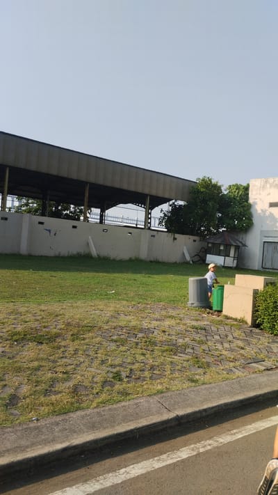 In Trosobo, Indonesia, an outdoor scene unfolds under a clear sky. A young boy wearing a light-colored cap and a striped shirt stands near two large waste bins, one gray and one green. He appears to be interacting with the bins or the area around them. Behind him, a low, gray concrete wall stretches across the frame, with a large, covered structure featuring a corrugated metal roof extending beyond it. Lush green grass covers the ground in front of the wall, with a section of patterned brickwork visible in the foreground. To the right of the boy, a small, weathered structure with a pointed roof sits nestled amongst trees. A portion of a white building with closed window shutters is visible to the far right. The scene appears to be set in an open, possibly industrial or recreational area.
