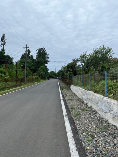 The image depicts a rural road in the village of Ghrmaghele Community, Georgia. The road is paved and has white lines marking the edges. On the left side of the road, there is a wire fence, utility poles with electrical wires, and dense vegetation including trees and bushes. On the right side of the road, there is a low concrete wall topped with a wire fence. Beyond the fence, there is more dense vegetation and a glimpse of a building. The sky is overcast with scattered clouds. Two cars are parked on the side of the road in the distance. The scene suggests a quiet, possibly overcast day in a semi-rural area.
