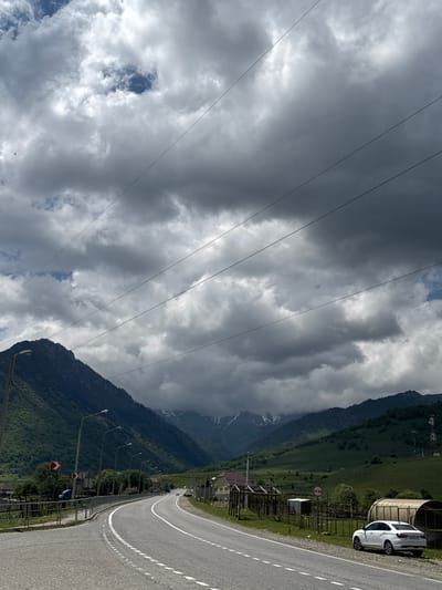 A white car is parked on the right side of a paved road that curves to the left and recedes into the distance. The road is bordered by a grassy shoulder and a fence with various structures behind it, including a large, covered cylindrical structure. Beyond the fence, green hills rise up to meet dark, imposing mountains, some of which are capped with snow. A telecommunications tower stands on the right side of the frame, further in the distance. Overhead, a dramatic sky is filled with dark, heavy clouds interspersed with patches of blue. The scene appears to be set in a rural or mountainous area.