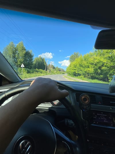 The image is taken from the driver's perspective inside a car, looking forward through the windshield. A person's hand is visible on the steering wheel. The car is on a paved road that curves to the right, surrounded by lush green trees on both sides. The sky is bright blue with some white clouds. Power lines are visible stretching across the sky. There are also some road signs, including a triangular warning sign on the left. In the distance, there are utility poles. The car's dashboard is partially visible, showing a speedometer and infotainment system display. The infotainment screen displays Russian text, indicating the car is likely in Russia. One of the texts reads "Долговременнo" which translates to "Long-term". The presence of Russian text on the car's infotainment system and the road signs suggest the location could be in or near Васильево, Russia.