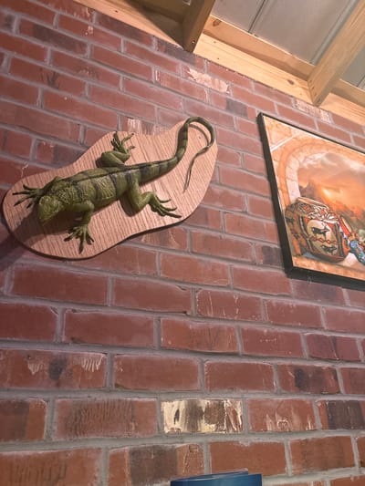 A mounted green lizard adorns a brick wall, positioned below a framed print featuring earthy tones and a central, stylized image. The lizard, seemingly taxidermied or sculpted, rests on an oval wooden plaque. A portion of exposed wood framing is visible on the left, along with a hint of a blue object on the right. The brick wall is textured with variations in color and mortar lines.