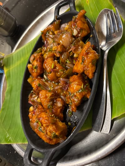 The image depicts a close-up of a serving of Chicken Manchurian, a popular Indo-Chinese dish, presented in a black oval serving dish. The dish rests on a vibrant green banana leaf, which in turn lies on a larger, silver-colored platter. A serving spoon and fork are placed beside the serving dish on the banana leaf. The Chicken Manchurian appears to consist of bite-sized pieces of fried chicken coated in a dark brown, slightly glossy sauce. Diced onions and green herbs are visible within the sauce. The image was taken in Kamalapura, India.