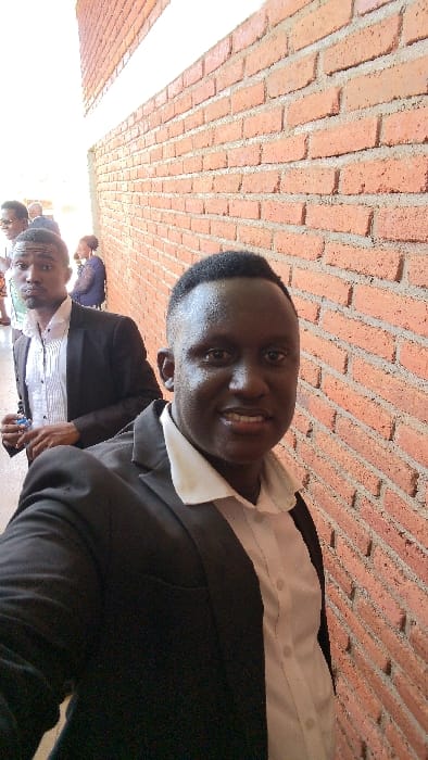 Two men in black suits and white shirts stand outside a brick building. The closer man smiles at the camera. A brick wall is prominently displayed, and other individuals are visible in the background, albeit blurry.