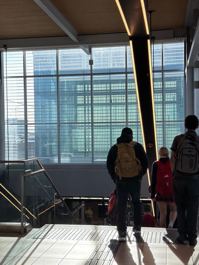 A vertically oriented photograph captures three individuals standing in a modern indoor public space, likely a transit station or concourse in Brossard, Canada. The setting features large grid-patterned windows on the left, allowing ample natural light to illuminate the scene, and a tiled floor with tactile paving on the right.
In the foreground, a person with their back to the viewer wears a dark jacket, black cap, and a tan/beige tactical backpack. Behind them, another individual is seen wearing a red top with a white Adidas logo and dark pants, with white socks visible. Further back, a third person in a grey and striped shirt carries a grey backpack with the word "SLUICE" visible on its surface. The individuals appear to be standing or waiting.
A prominent dark horizontal beam with warm-toned LED strip lighting runs across the middle ground. Strong, elongated shadows cast on the tiled floor by the people and architectural elements indicate bright, directional sunlight, suggesting it is late morning or afternoon on a clear day. Reflections of the interior structure are visible in the windows. A modern glass and metal railing of a staircase is partially visible in the lower right corner.