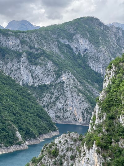 This is a landscape image of a canyon with steep cliffs and a body of water at the bottom. The cliffs are predominantly gray rock with varying amounts of green vegetation clinging to the surfaces. The water is a deep blue color, likely a lake or river. A mountain peak is visible in the distance on the left side of the image, partially obscured by clouds. This location is most likely the Piva Canyon in Plužine, Montenegro.
