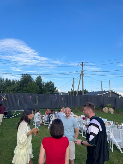 A group of people are gathered in a backyard in Simagino, Russia, for an outdoor event. The scene is set on a well-maintained lawn with a black fence in the background, separating the yard from a line of trees and houses. A long table, covered with a white tablecloth, is laden with food and set with white chairs, indicating a meal or celebration. Several individuals are seen interacting around the table and in the foreground. In the foreground, a woman with long dark hair wearing a light yellow dress is holding a drink and a small green object. She is facing away from the camera, looking towards another person. To her right, a man with short blonde hair wearing a light blue t-shirt and beige shorts is smiling and holding a small plate. Next to him, a man in a black and white athletic jacket is facing to the left and appears to be talking. The sky above is a clear blue with some wispy white clouds, and utility poles with numerous wires are visible against the sky. Decorative ribbons in various colors are attached to the fence. The overall atmosphere suggests a relaxed social gathering on a pleasant day.