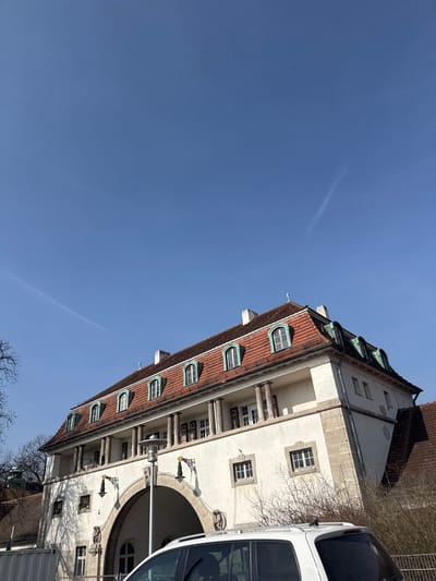 The image displays an upward-angled view of a historical architectural structure, specifically one of the bathhouse pavilions of the Sprudelhof complex in Bad Nauheim, Germany, under a clear, bright blue sky with faint aircraft contrails.
The prominent building features a red tiled hip roof with numerous arched dormer windows, each framed in a light green or patinated copper color with white sashes. Several white chimneys rise from the roofline. Below the roof is a white stucco facade. The upper section of the building includes a loggia or covered balcony supported by multiple cylindrical columns. Behind these columns are windows and doors with dark frames.
At the mid-level, square windows with grid patterns are visible. The ground level features a large, central arched entrance. Flanking this archway are decorative elements, including carved stone figures and ornate wall-mounted light fixtures with yellow accents, casting shadows on the facade. The building's corners are marked by rusticated stone blocks.
In the lower foreground, a partial view of a white vehicle, likely an SUV or van, obscures the bottom portion of the building. Only its roof, roof rails, and a segment of its rear window are visible. A tall, grey metallic lamppost with a circular fixture stands in front of the building, positioned to the left of the central archway. Bare tree branches are present on the left side of the frame, and leafless bushes are visible on the right, suggesting a dormant vegetative season. Strong sunlight illuminates the scene, creating defined shadows.