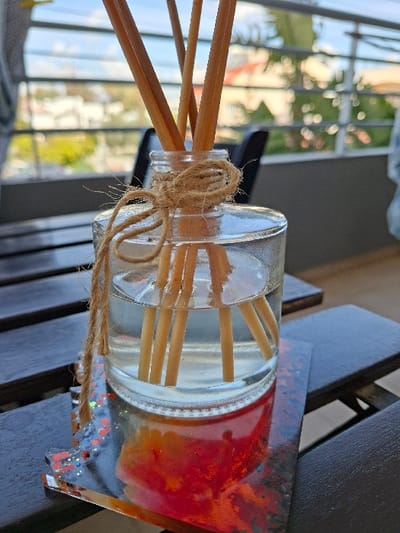 A clear glass diffuser bottle, approximately cylindrical with rounded shoulders, sits on a multi-colored coaster featuring abstract patterns of red, orange, and black, with sparse iridescent flecks. The bottle contains a clear, translucent liquid and approximately ten to twelve light brown wooden reeds extending upward from the liquid. A piece of light brown jute twine is wrapped around the neck of the bottle, tied in a knot with a loose end hanging down the side. This arrangement rests on a dark, slatted wooden surface, likely an outdoor table or balcony railing. The background, out of focus, reveals an outdoor environment in Agios Dometios, Cyprus, characterized by a light-colored railing, distant green foliage, blurred urban structures, and a clear blue sky, indicative of daytime conditions. No individuals or overt actions are depicted.