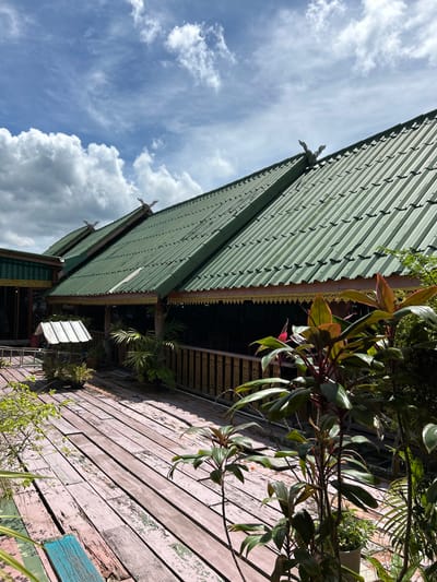 The image captures a view of the exterior of a traditional building, likely a restaurant or guesthouse, located in Tambon Ko Panyi, Thailand. The building features a prominent green corrugated metal roof with ornate decorative elements along the eaves and at the peak of the gables. A wooden walkway, weathered and painted in shades of pink and blue, extends across the foreground. Various potted plants, including those with large, glossy leaves and vibrant red stems, are placed on the walkway and along the railing of the building's veranda. A small, white structure resembling a well or information booth stands on the left side of the walkway. The background reveals a cloudy blue sky, suggesting daytime. The overall scene depicts a tropical, possibly waterfront, setting with traditional architecture and lush vegetation. There are no people or discernible actions in the image.