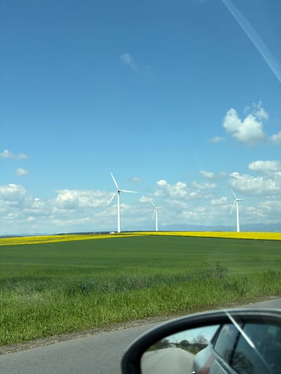 This media file captures a bright, sunny daytime scene of a rural landscape, likely taken from a moving vehicle in Chargan, Bulgaria. The sky dominates the upper portion of the image, showcasing a clear blue expanse dotted with scattered white cumulus clouds. A faint, linear contrail from an aircraft is visible in the upper left section of the sky.

In the midground, several prominent white wind turbines with three blades stand tall against the horizon, signifying a modern energy landscape. These turbines are spaced out across the vista, adding to the sense of open space.

Below the horizon, expansive agricultural fields stretch into the distance. A striking feature is a broad strip of vibrant yellow flowering crops, characteristic of rapeseed or canola in bloom, running parallel to the road. This yellow field contrasts with a wider area of lush green crops or grass closer to the foreground.

A paved road runs along the right side of the image, with a grassy verge separating it from the fields. The photograph is taken from the perspective of a vehicle, as evidenced by the car's side mirror visible in the far right foreground. The mirror's reflection shows trees and possibly the road extending behind the vehicle.

The scene conveys a sense of peaceful, open countryside under a clear sky, typical of late spring or early summer. There is no visible text in the image.